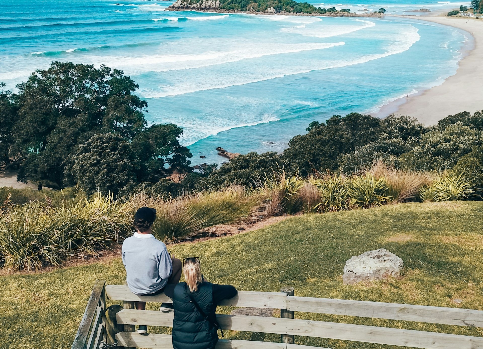 Two people stand at a wooden railing surrounded by greenery, overlooking a scenic beach with waves on Chatham Island. It's a clear day.