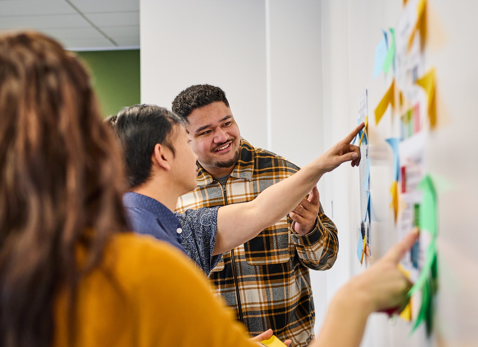 Three people are standing, sticking post-it notes to a wall. The post-it notes have writing on them.